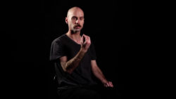 nathan walker, bald moustachioed man sitting in dark t-shirt in dark stage, holding hand and fingers in front of face