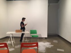 kimberley standing in gallery room reading from paper. Table and coloured chairs and scattered papers on ground