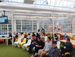 group of phd students on chairs in large open room with green carpets and glass windows all around like a large greenhouse.