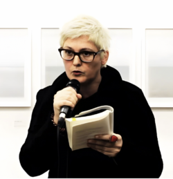 woman with white short hair and glasses, reads poetry into a microphone